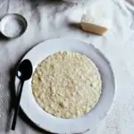 A top down photo of a white plate containing cacio e pepe risotto. A black spoon rests on the edge of the plate, and on the table there is a block of pecorino romano cheese, and a small bowl of salt.