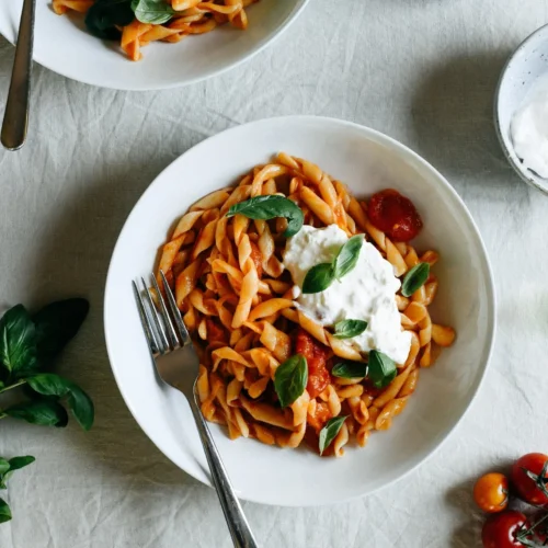 A flat lay image of two bowls of pasta, containing a hand formed spiral shape called busiate and dressed in a fresh cherry tomato sauce. Topped with burrata cheese and fresh basil leaves.