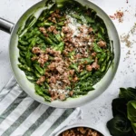 A top down photo of a large aluminium sauté pan filled with a garganelli pasta, coated in a vibrant green spinach sauce topped with crispy sausage pieces and grated Parmigiano Reggiano cheese.