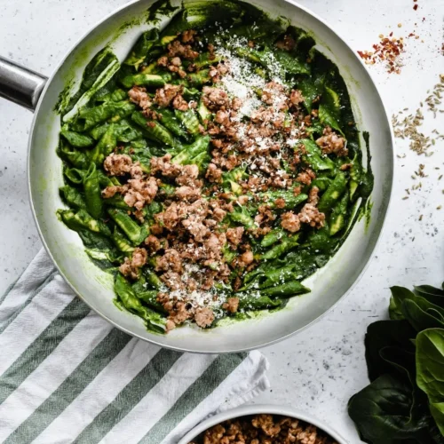 A top down photo of a large aluminium sauté pan filled with a garganelli pasta, coated in a vibrant green spinach sauce topped with crispy sausage pieces and grated Parmigiano Reggiano cheese.