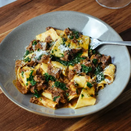 A bowl of fresh pappardelle pasta with ossobuco sauce, garnished with a vibrant green gremolata.