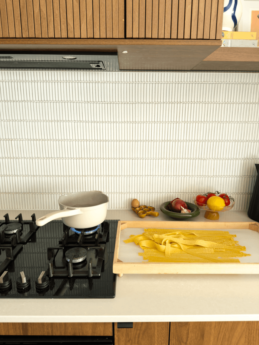 A kitchen counter is shown with a saucepan of water coming to a boil on a stove top. Next to the stove top a mesh drying rack with pappardelle pasta lying on it is visible. 