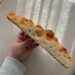 A slice of focaccia is held displaying the crumb, featuring large air pockets and a crisp, golden exterior.