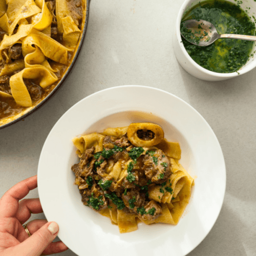 A top-down view of a white bistro plate containing a classic ossobuco ragu, served with fresh pappardelle and a vibrant green gremolata.