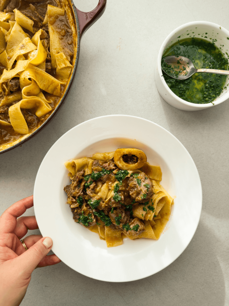 A top-down view of a white bistro plate containing ossobuco ragu, served with fresh pappardelle and a vibrant green gremolata.