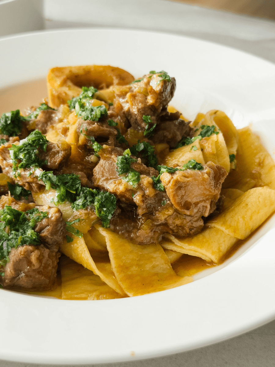 A close up shot of a plate of ossobucco ragu, served with fresh egg pasta and gremolata sauce. A bone with marrow in its centre is visible in the back of the image, a common way to serve ossobuco. 