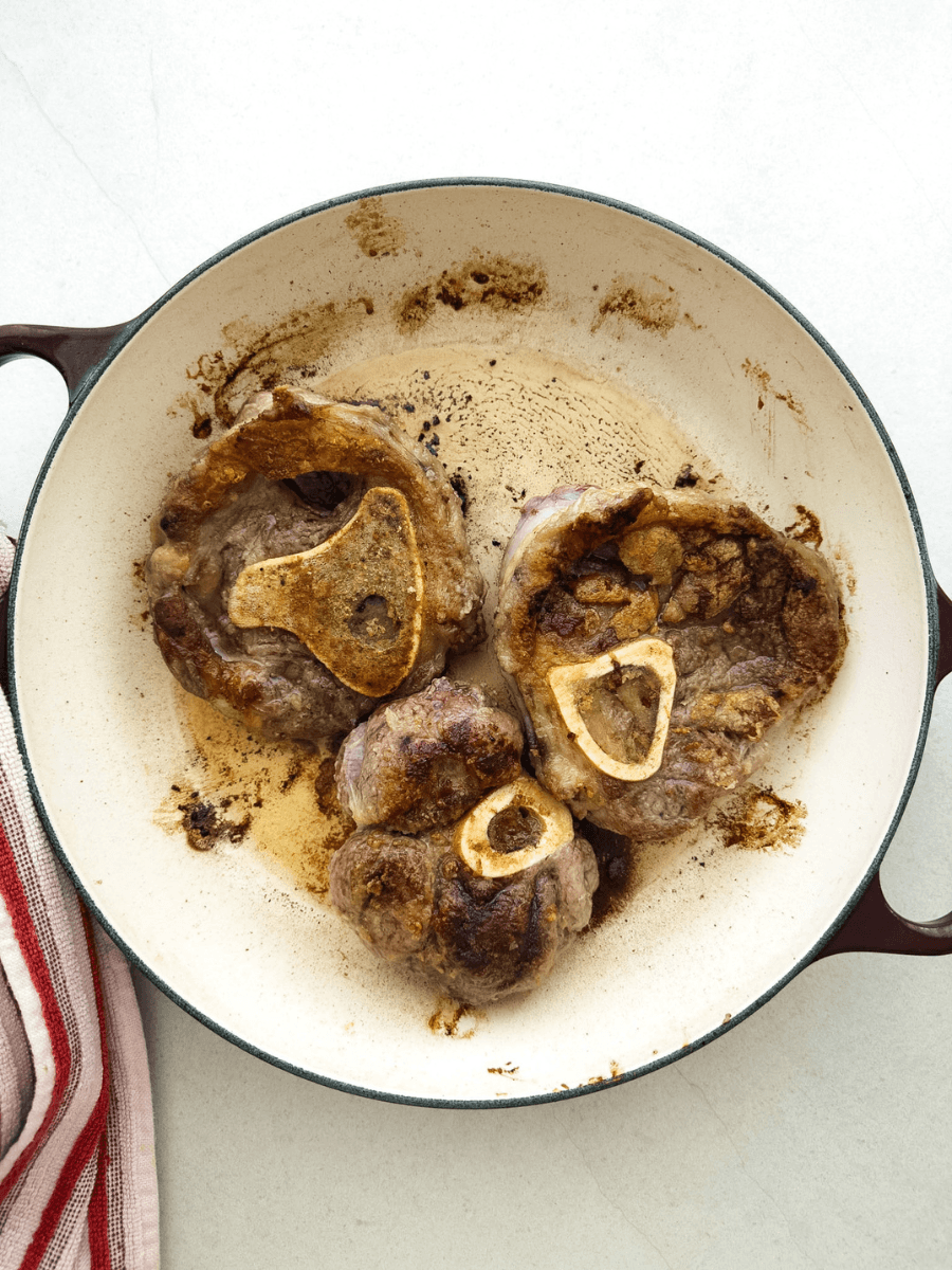 Seared ossobuco pieces with a dark brown crust are shown top-down in a large heavy-based pan. 