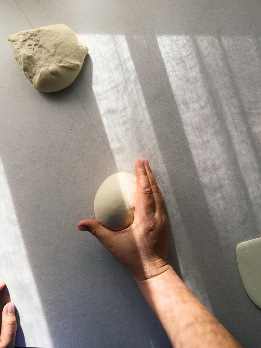 A top down view of a hand in motion shaping a dough ball. The hand is cupped around the ball in the shape of a c to create tension. 