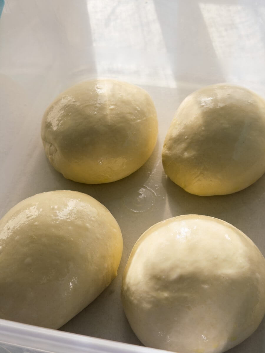 Prehshaped pizza dough balls brushed lightly in olive oil sit inside a large Tupperware container. 