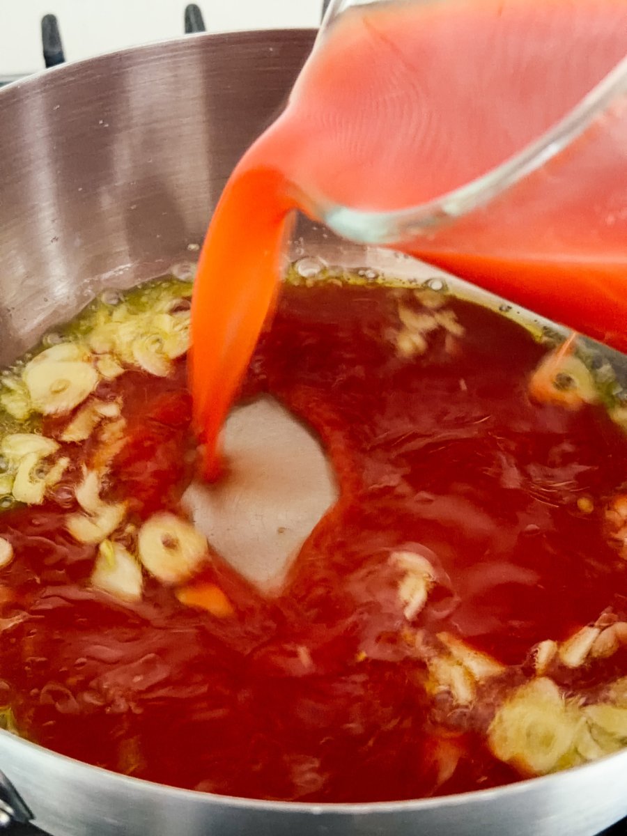 A measuring jug pours in a tomato paste and water mixture to a pan of garlic. 