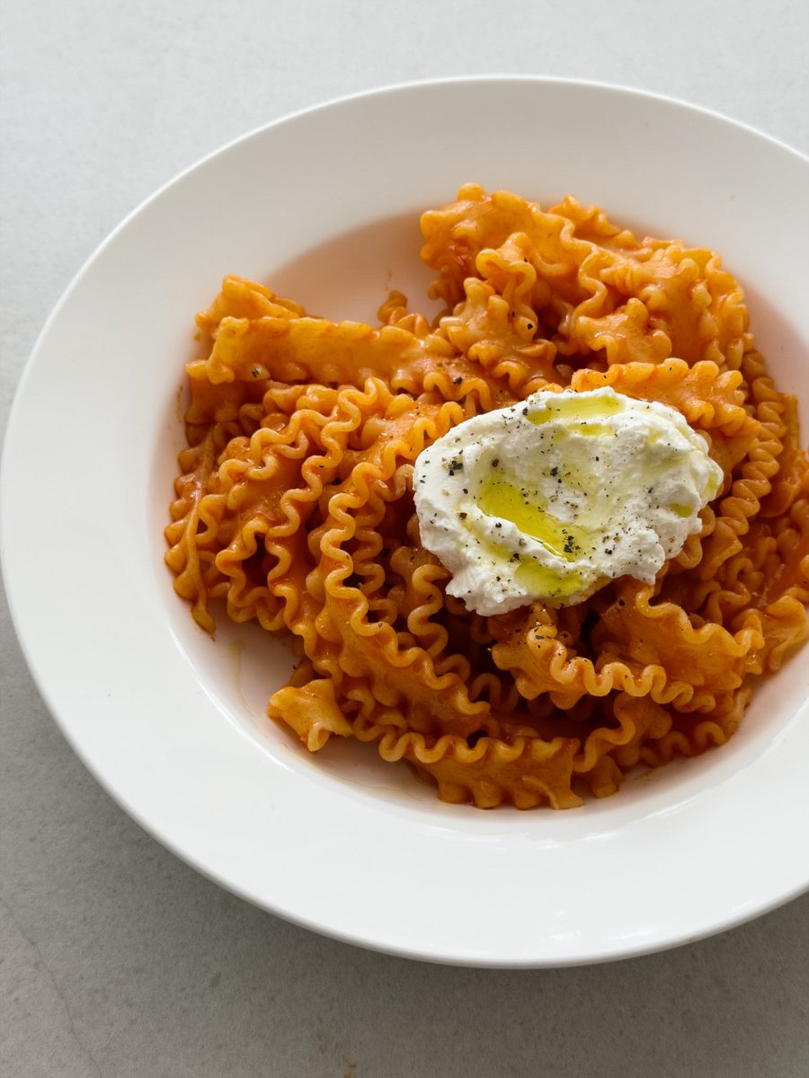 A close up shot of a white pasta bowl containing mafalde pasta, coated in a rich tomato sauce and served with a dollop of ricotta cheese, a drizzle of olive oil and a crack of black pepper. 