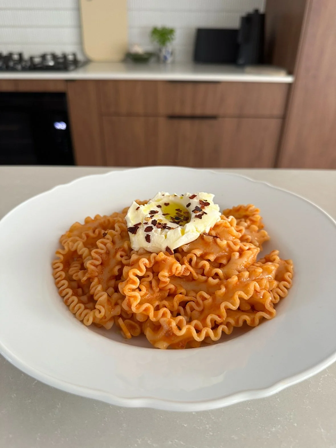 A photo of a white pasta plate filled with curl pappardelle pasta, coated in a simple tomato paste pasta sauce, served with a dollop of ricotta cheese and chilli flakes.