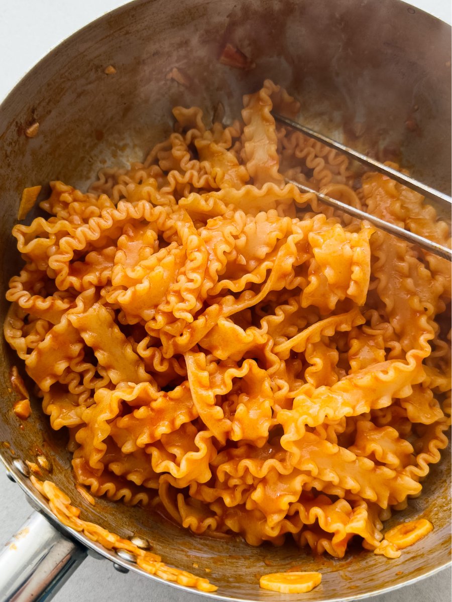A stainless steel pan containing curly pappardelle pasta, coated in a rich tomato paste pasta sauce. 