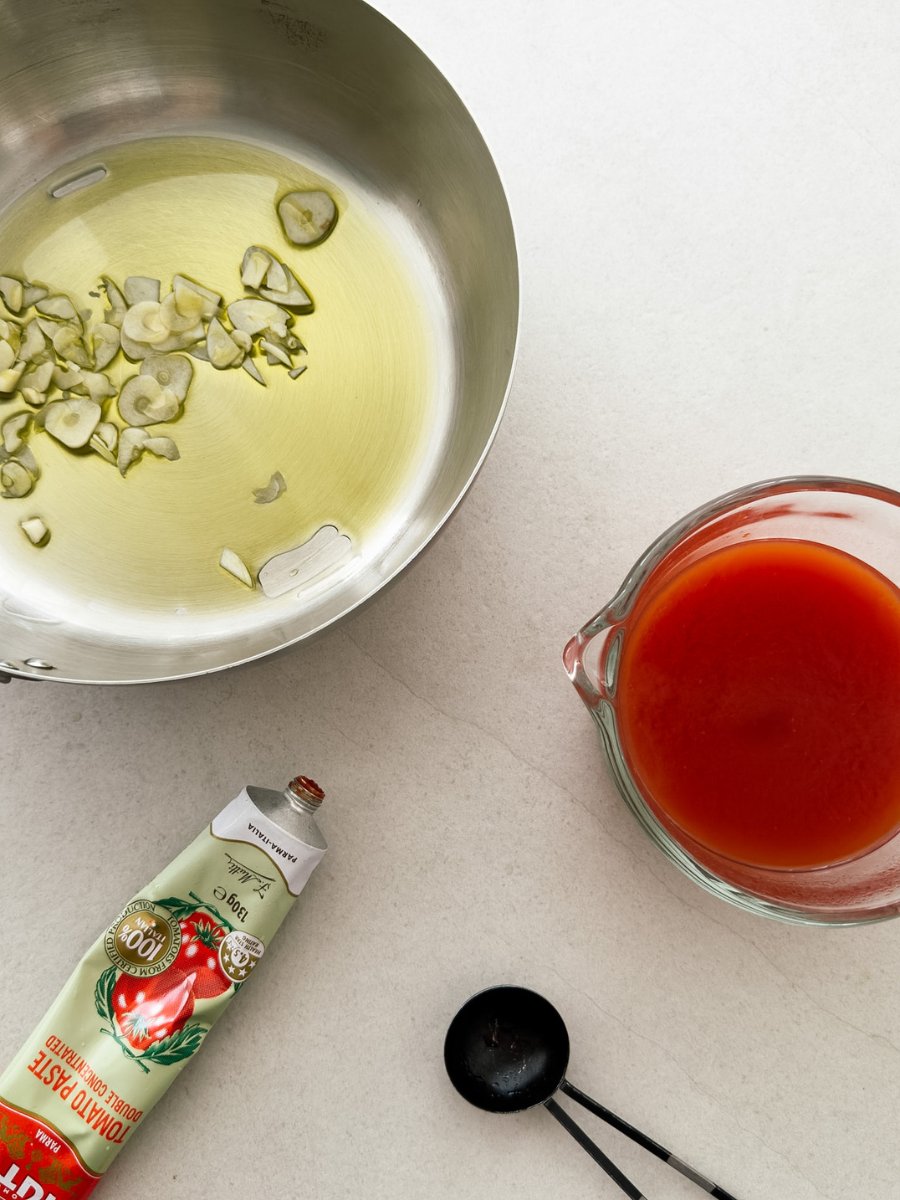 A sauteuse pan with olive oil and thinly sliced garlic cloves appears next to an open tube of tomato paste, and a measuring jug filled with a tomato paste dilution. 