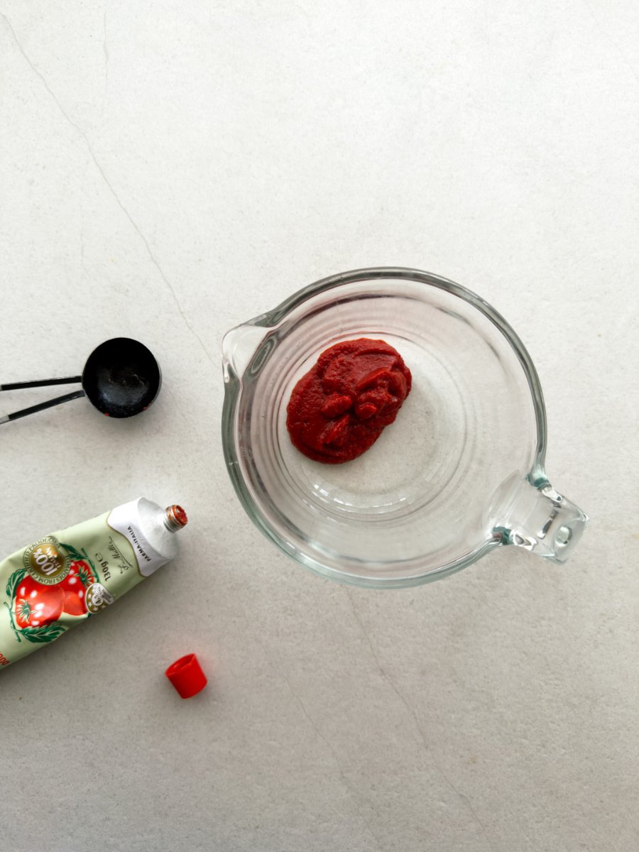 A top down shot of an open tube of tomato paste, measuring spoon and a jug with tomato paste inside. 