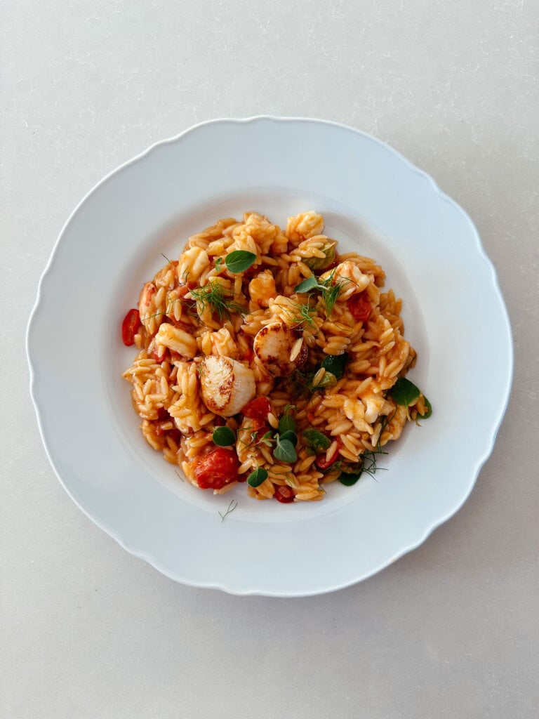 A top down photo of a white, scalloped edged bistro plate with a vibrant seafood orzo pasta, made with prawns, scallops, cherry tomatoes, fresh oregano and red chilli.