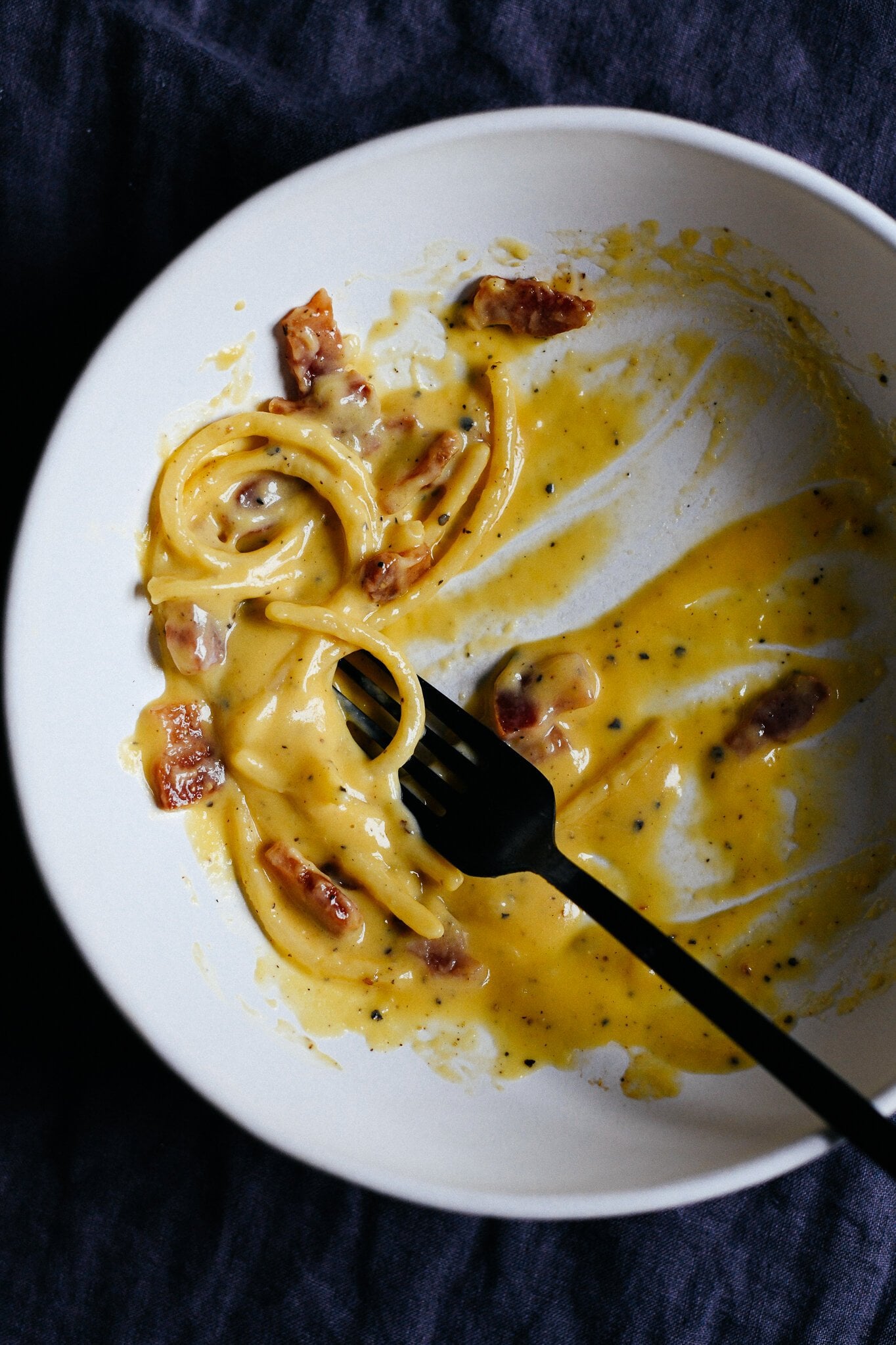 An almost finished bowl of Roman-style carbonara, with a creamy, silky sauce and just a few strands of spaghetti, and pieces of crispy guanciale remaining.