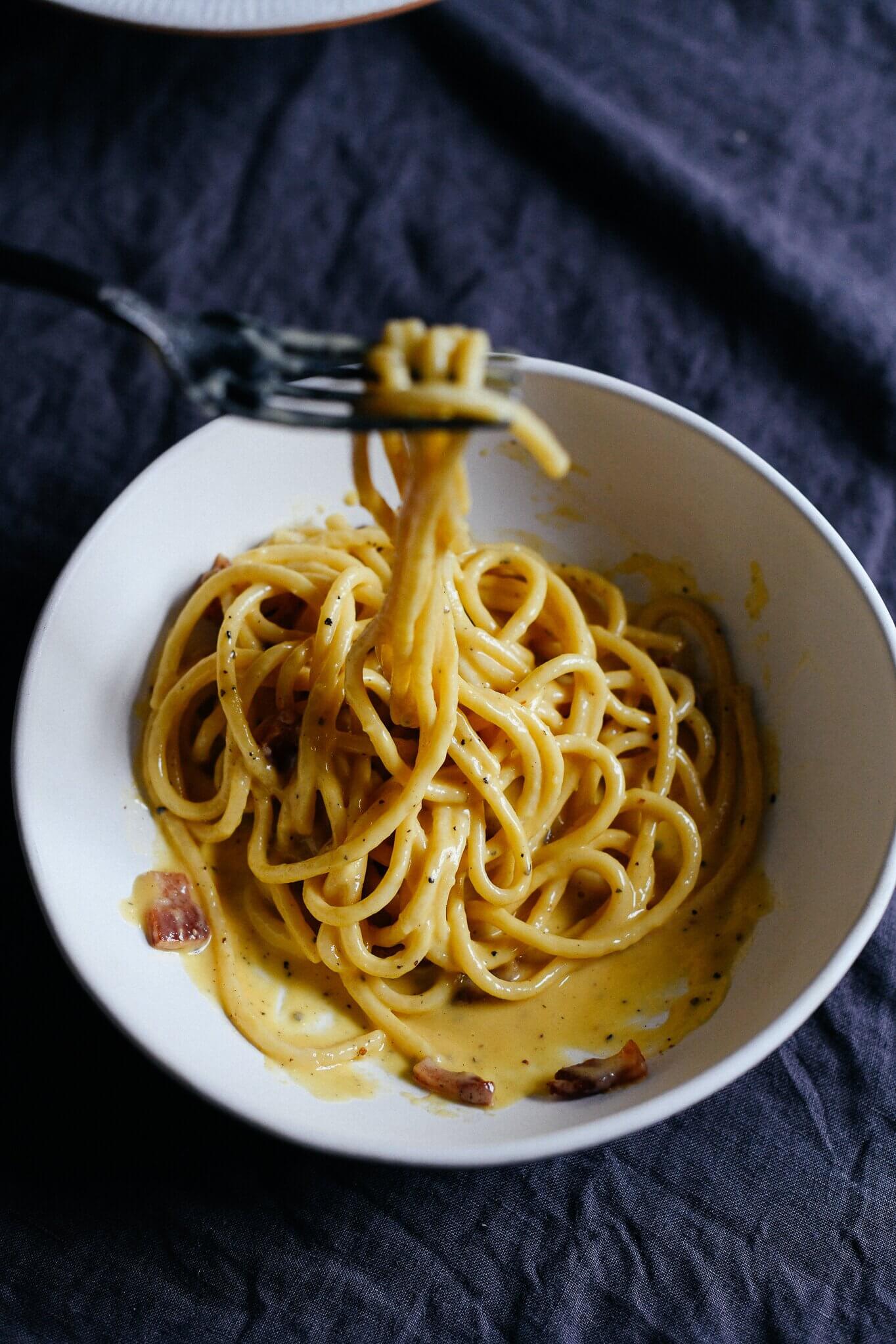 A close-up, top-down shot of authentic Roman-style carbonara pasta served in a shallow ceramic bowl. The glossy, creamy egg and Pecorino Romano sauce coats thick strands of spaghetti, garnished with crispy, golden-brown cubes of guanciale and a generous dusting of freshly cracked black pepper.