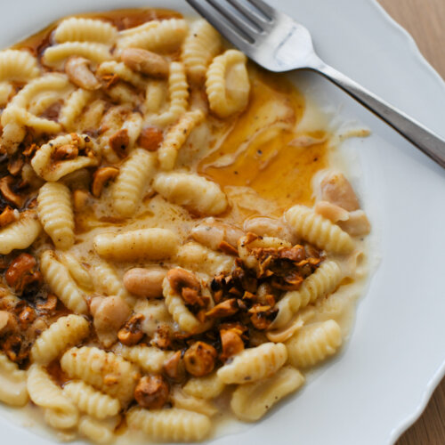 A white bistro plate with partially eaten, handmade cavatelli pasta in a creamy gorgonzola and white bean sauce, topped with brown butter and roasted hazelnuts.