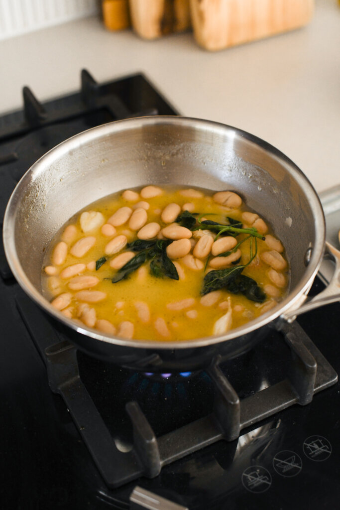 White beans simmering with sage and garlic in a shallow pan over a gas stove