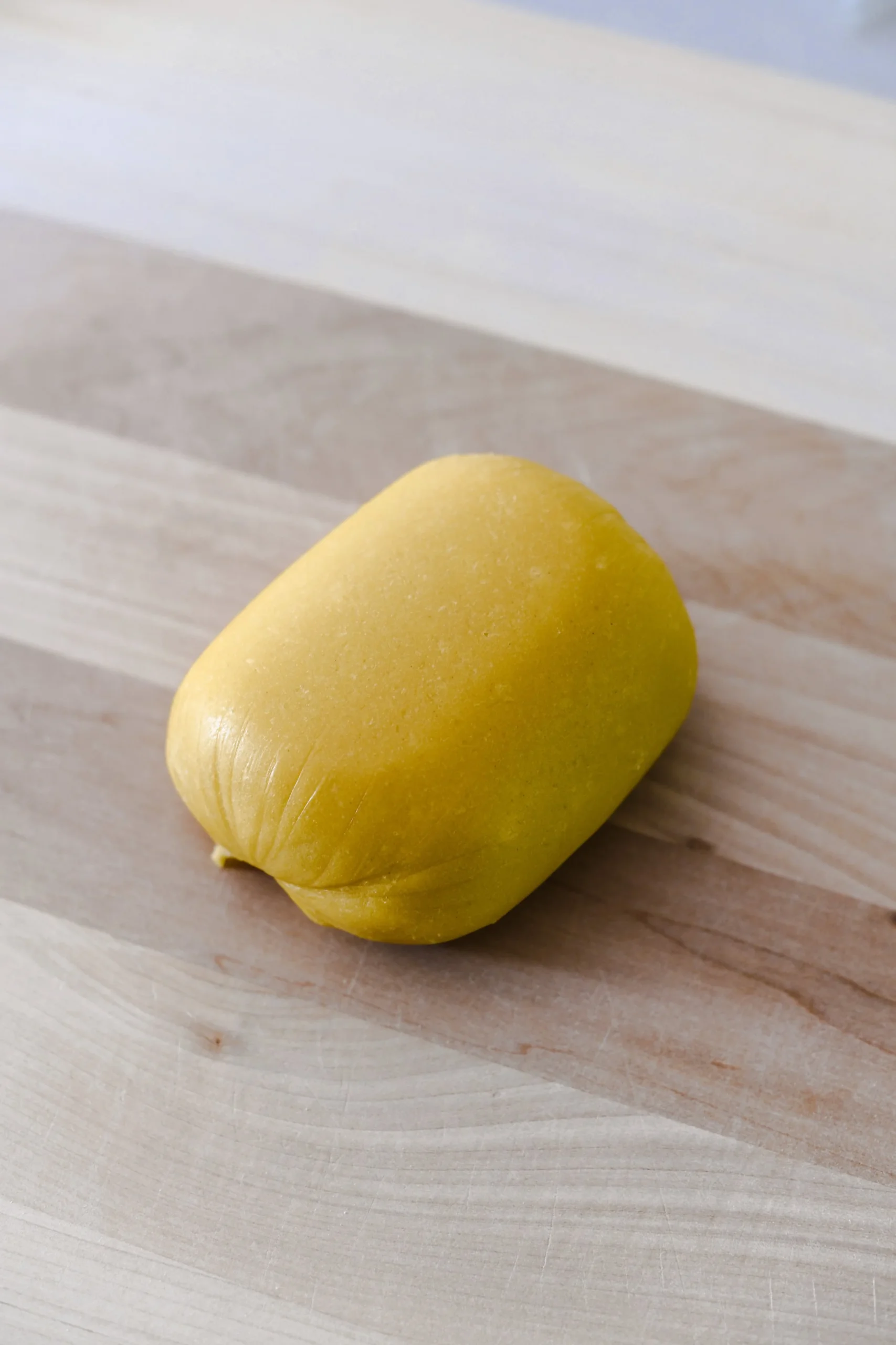 A smooth ball of fresh egg pasta dough sits on a wooden bench top. Visible air pockets can be seen beneath the surface layer of the pasta.