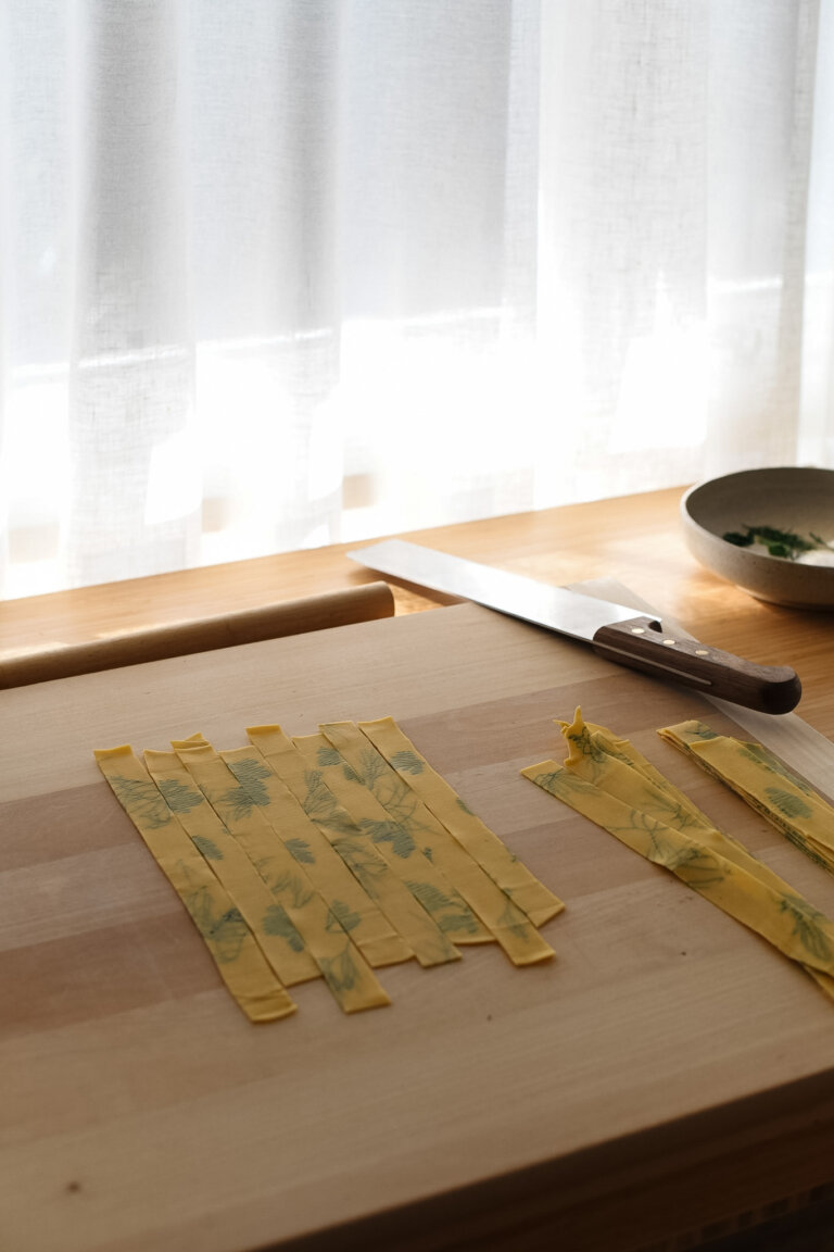 A work bench set up with a wooden pasta board and ribbon cut strands of herb laminated pasta placed neatly on top.