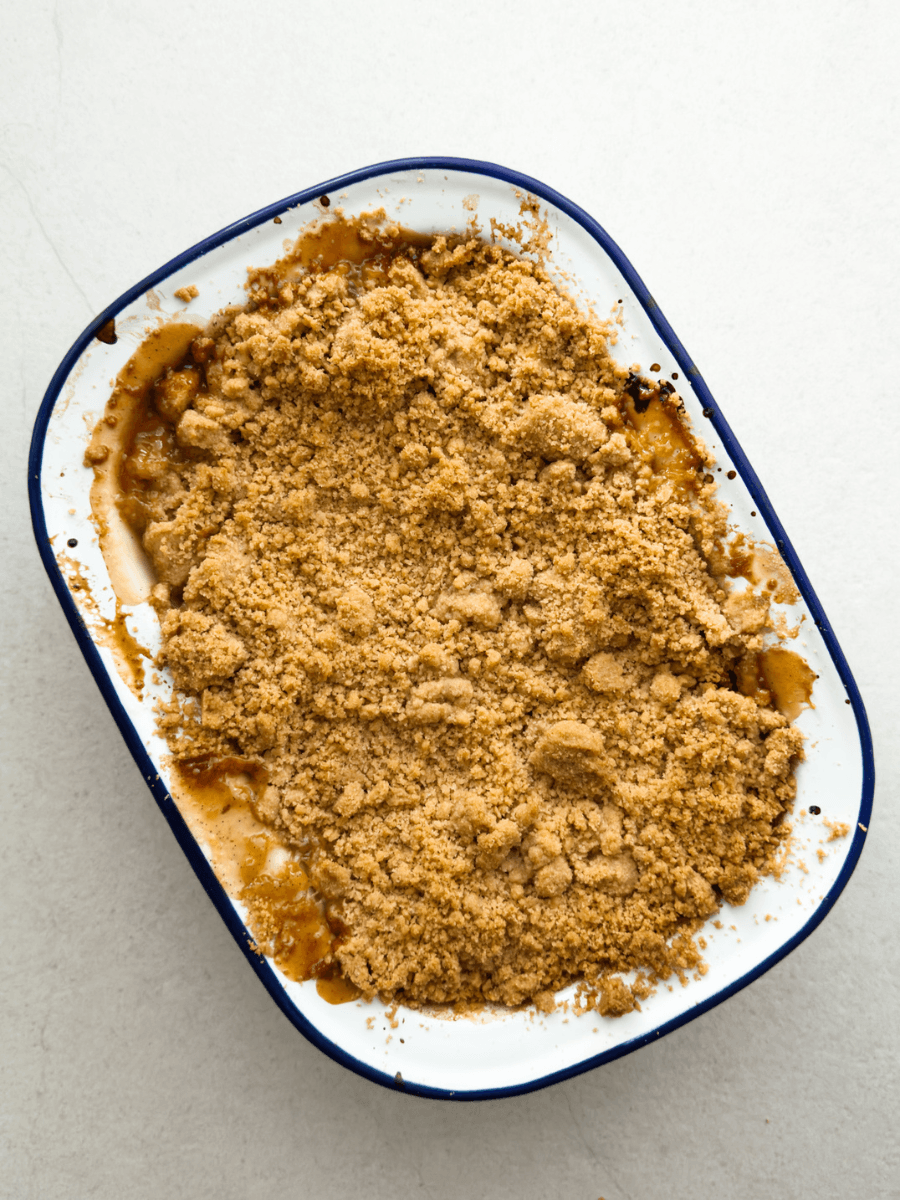 A freshly baked apple crumble is shown top-down, with a golden, crispy crumble topping and caramelised, bubbling apple filling at the edges. 