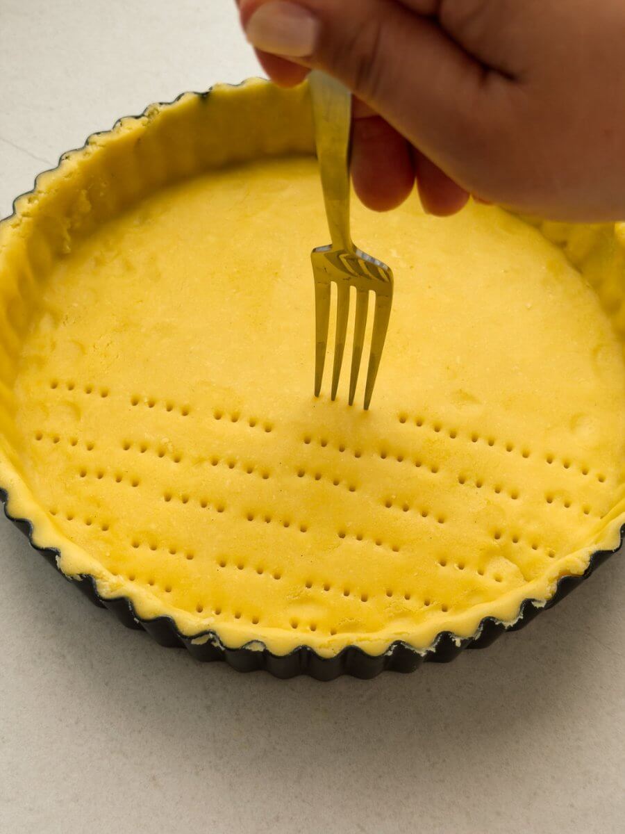 A hand holding a fork docks the base of a tart pan lined with a sweet pastry dough.