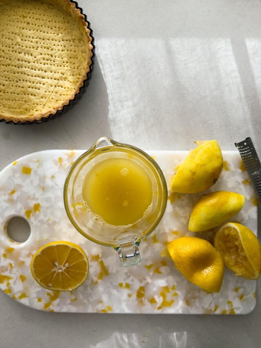 A top down shot of a yellow and white speckled chopping board, with a few Meyer lemon halves that have been juiced. A glass measuring jug filled with Meyer lemon juice sits next to the lemons, and a blind baked tart shell is in view.