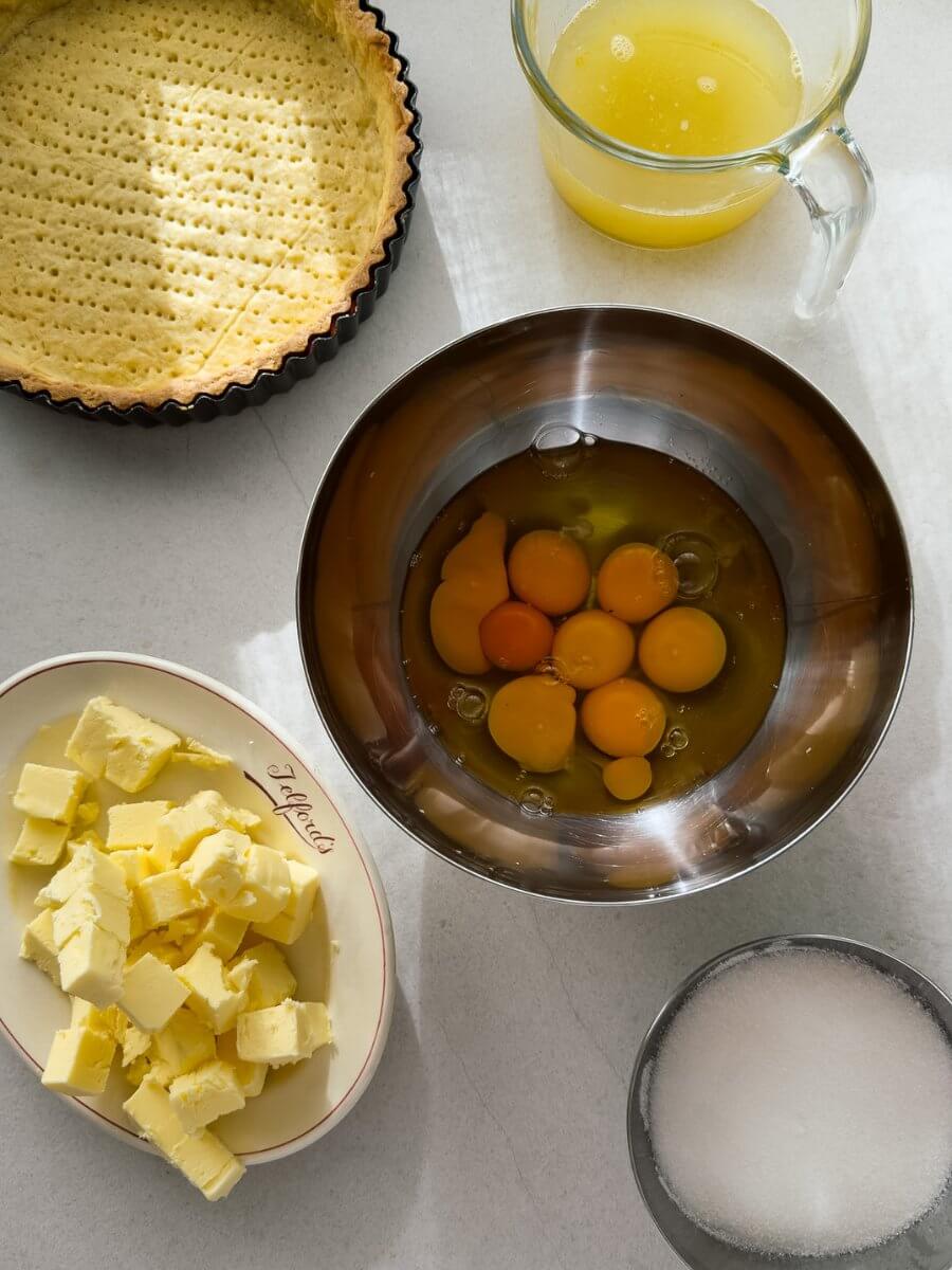 Flat-lay photo of the ingredients needed to make a classic lemon tart. A blind baked sweet pastry shell, lemon juice, a bowl of eggs and egg yolks, cubed butter and sugar.