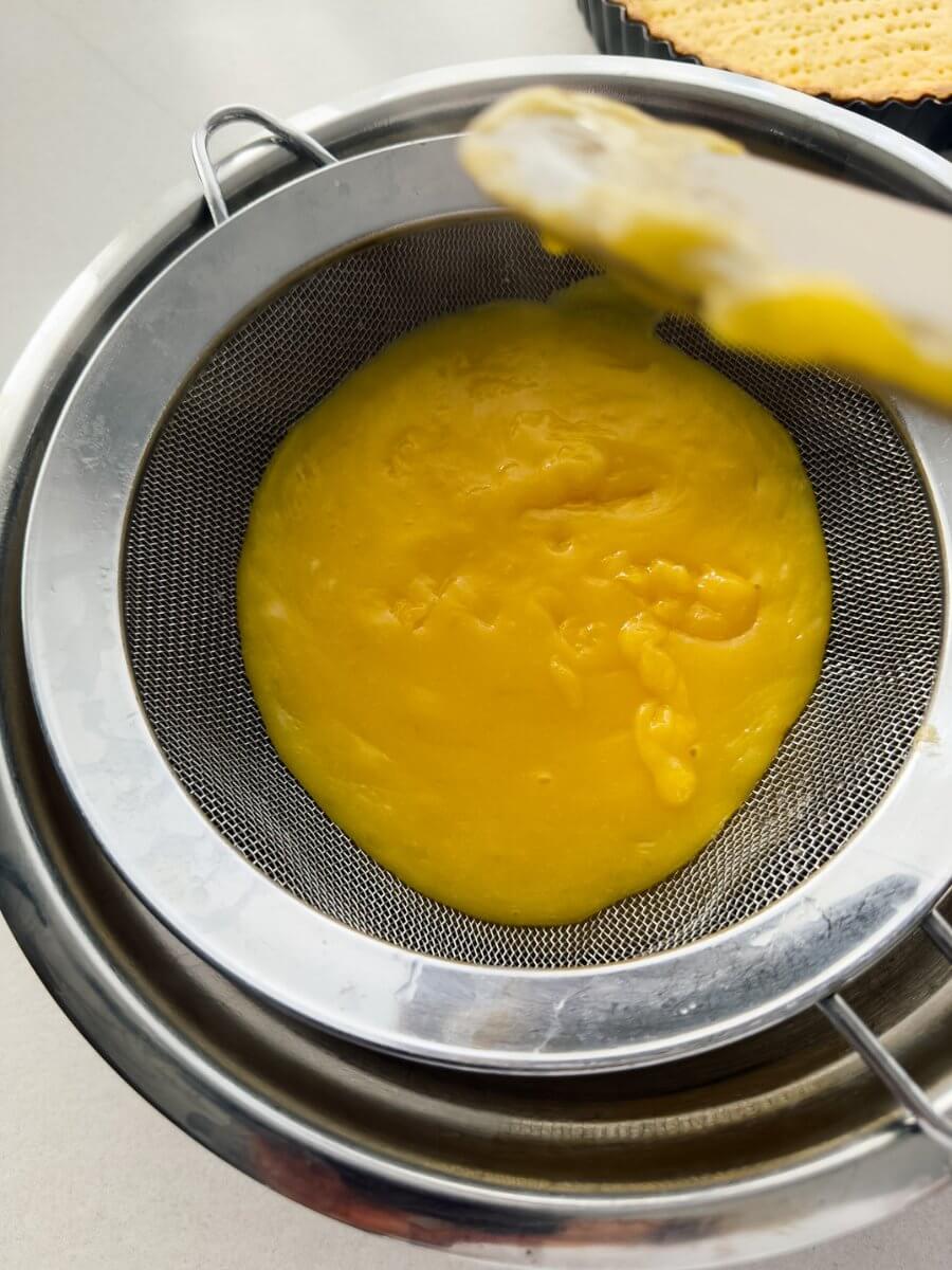 A top down photo showing freshly made lemon curd being passed through a sieve set over a bowl.