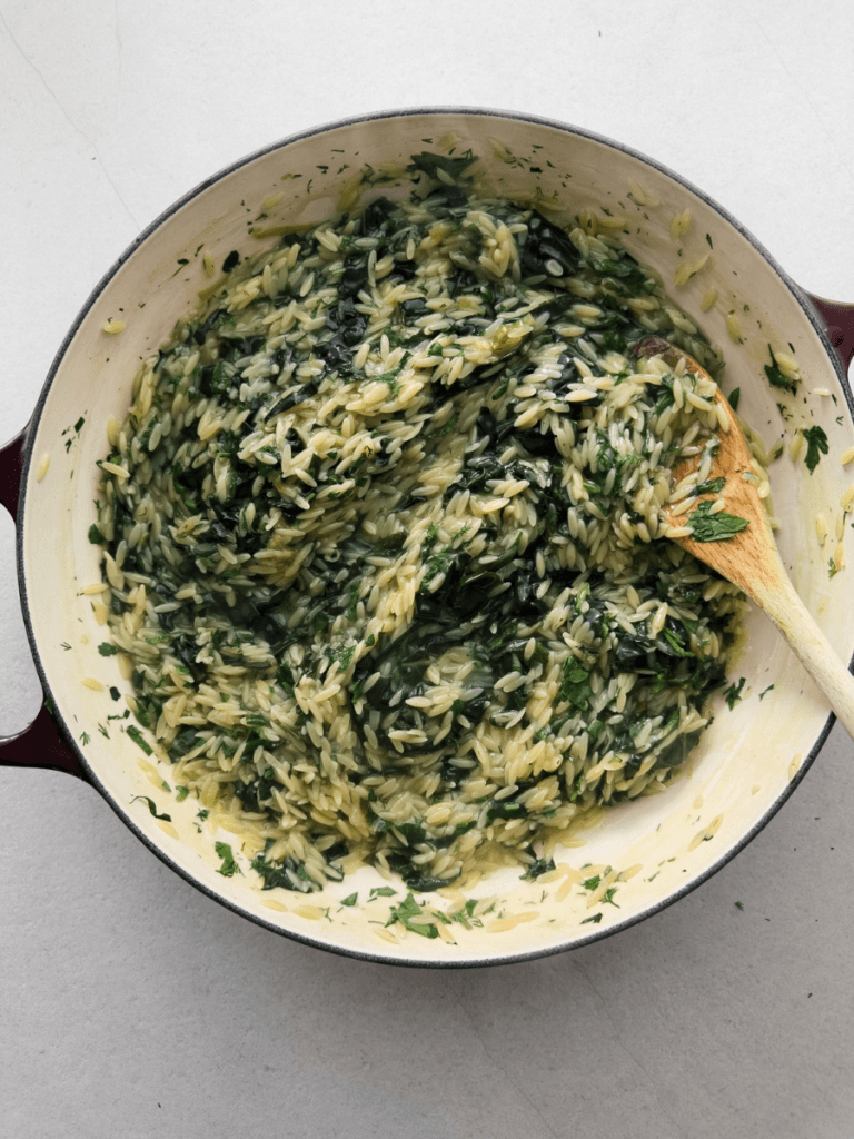 A top-down photo of a large skillet pan with cooked orzo pasta, silverbeet and herbs, making the perfect one pot spanakopita pasta.