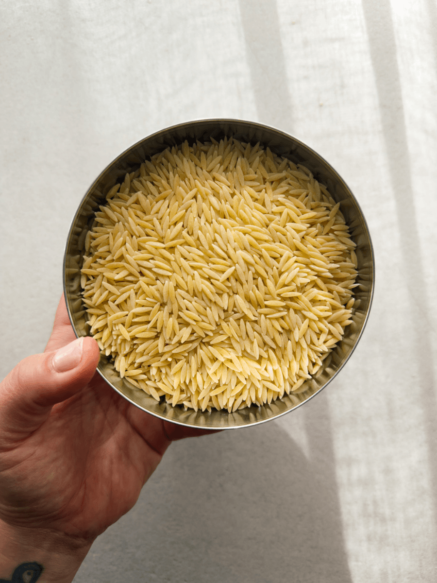 A small stainless steel bowl filled with uncooked, dry orzo pasta is held by a hand in the corner of the frame.