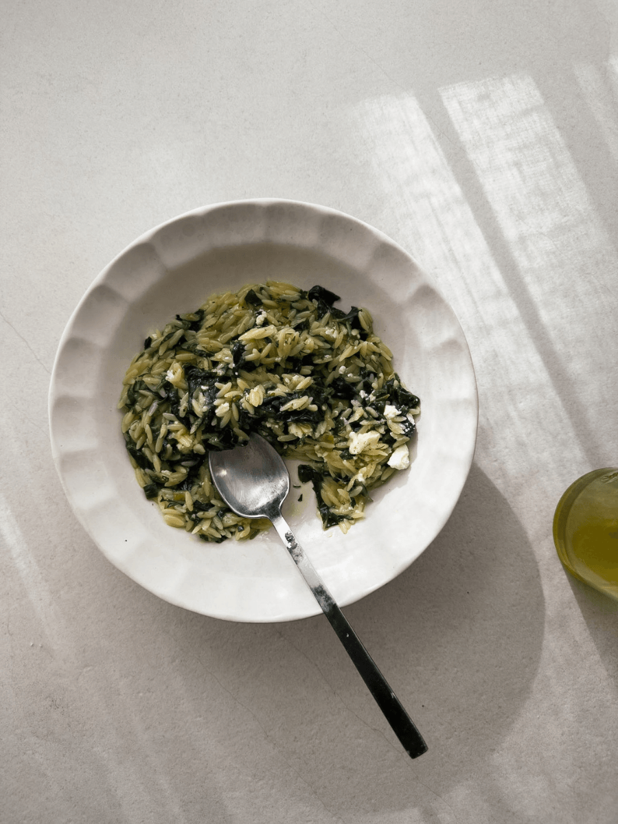 A top-down photo of a white bistro bowl with a detailed scalloped rim, filled with orzo pasta, silverbeet, herbs and topped with crumbs of Greek feta.