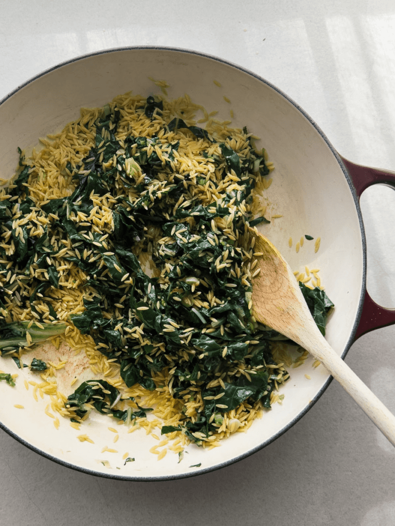 A top-down picture of a one pot spanakopita pasta - showing a large skillet pan with wilted silverbeet, spring onion greens and dry orzo pasta.