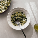 Orzo pasta with silverbeet, herbs and feta is shown in a white pasta bowl with scalloped edges. A casserole from which the pasta dish is served from is viewable in the top right corner.