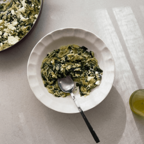 Orzo pasta with silverbeet, herbs and feta is shown in a white pasta bowl with scalloped edges. A casserole from which the pasta dish is served from is viewable in the top right corner.