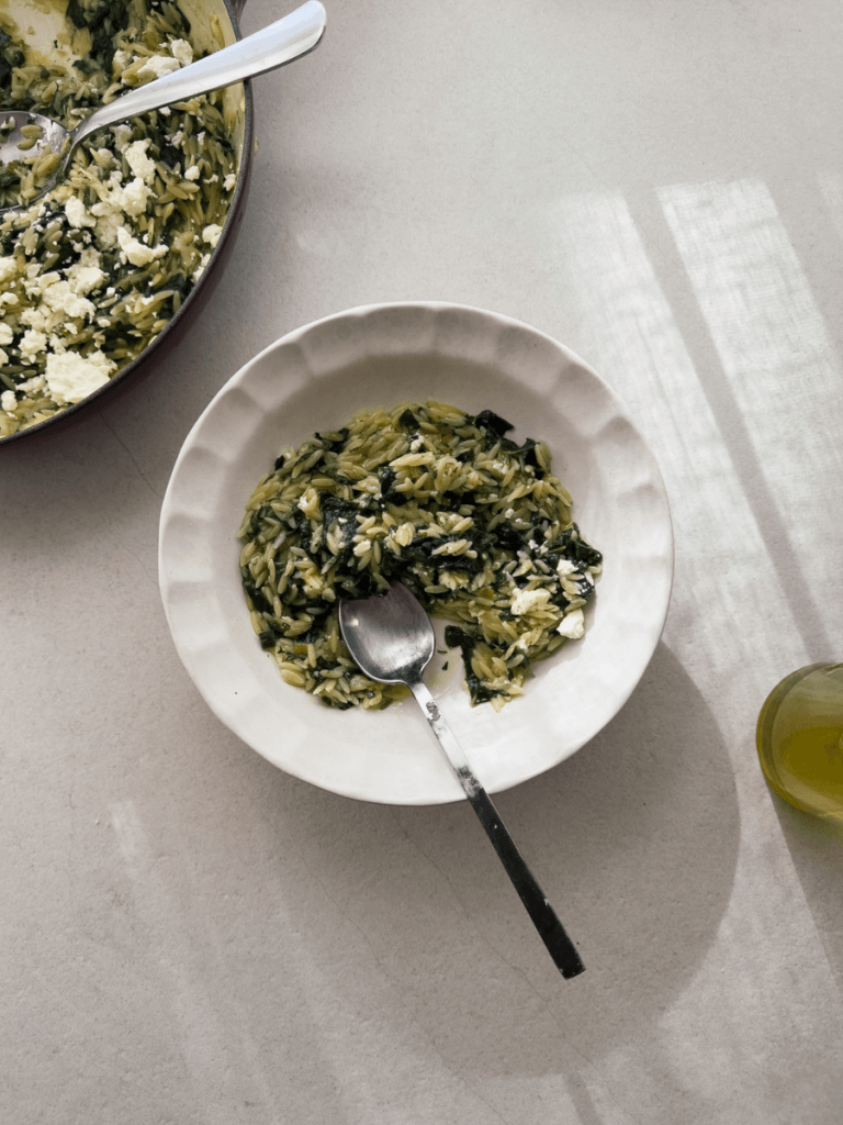 Orzo pasta with silverbeet, herbs and feta is shown in a white pasta bowl with scalloped edges. A casserole from which the pasta dish is served from is viewable in the top right corner.