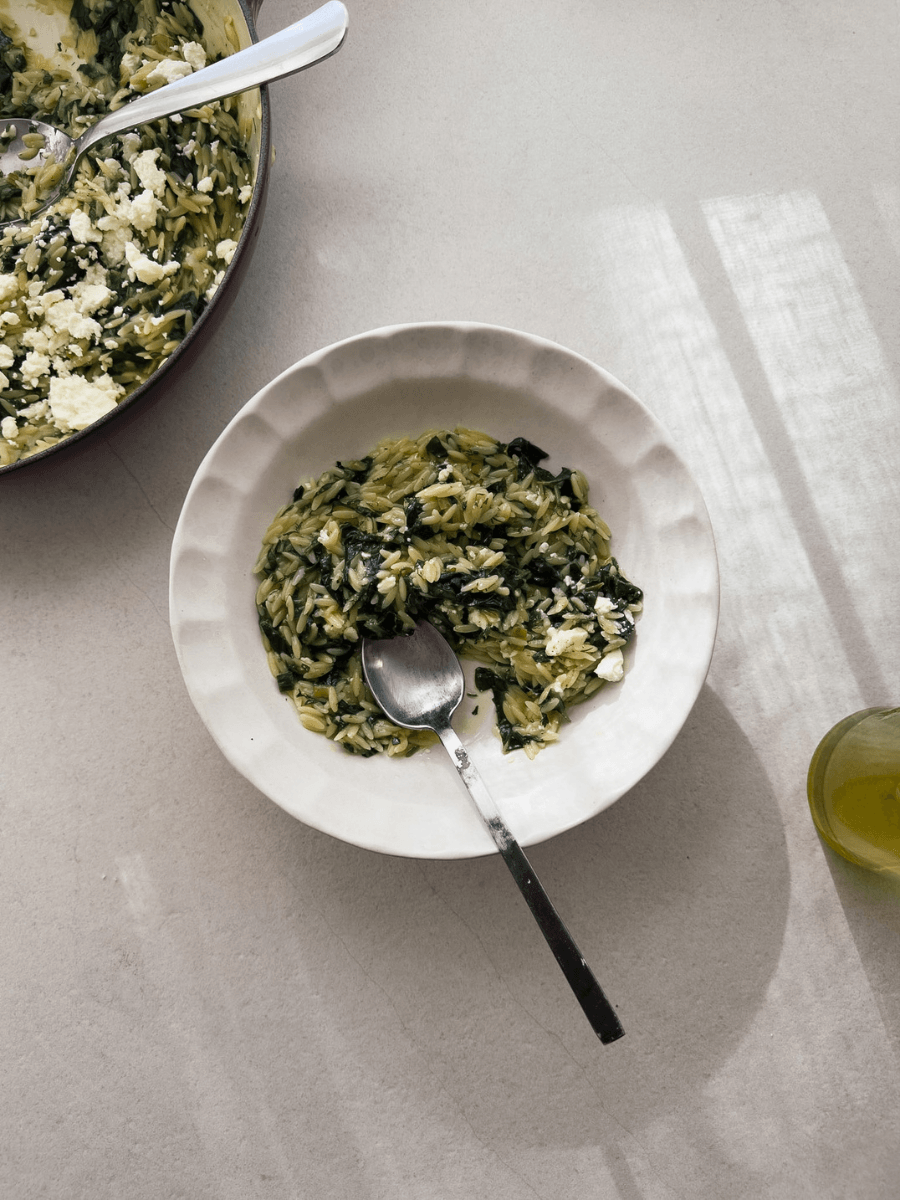 Orzo pasta with silverbeet, herbs and feta is shown in a white pasta bowl with scalloped edges. A casserole from which the pasta dish is served from is viewable in the top right corner.