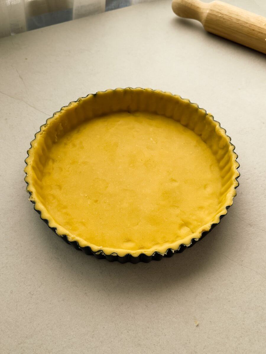 Trimmed sweet pastry dough lined a fluted tart pan. A rolling pin is visible in the backdrop.