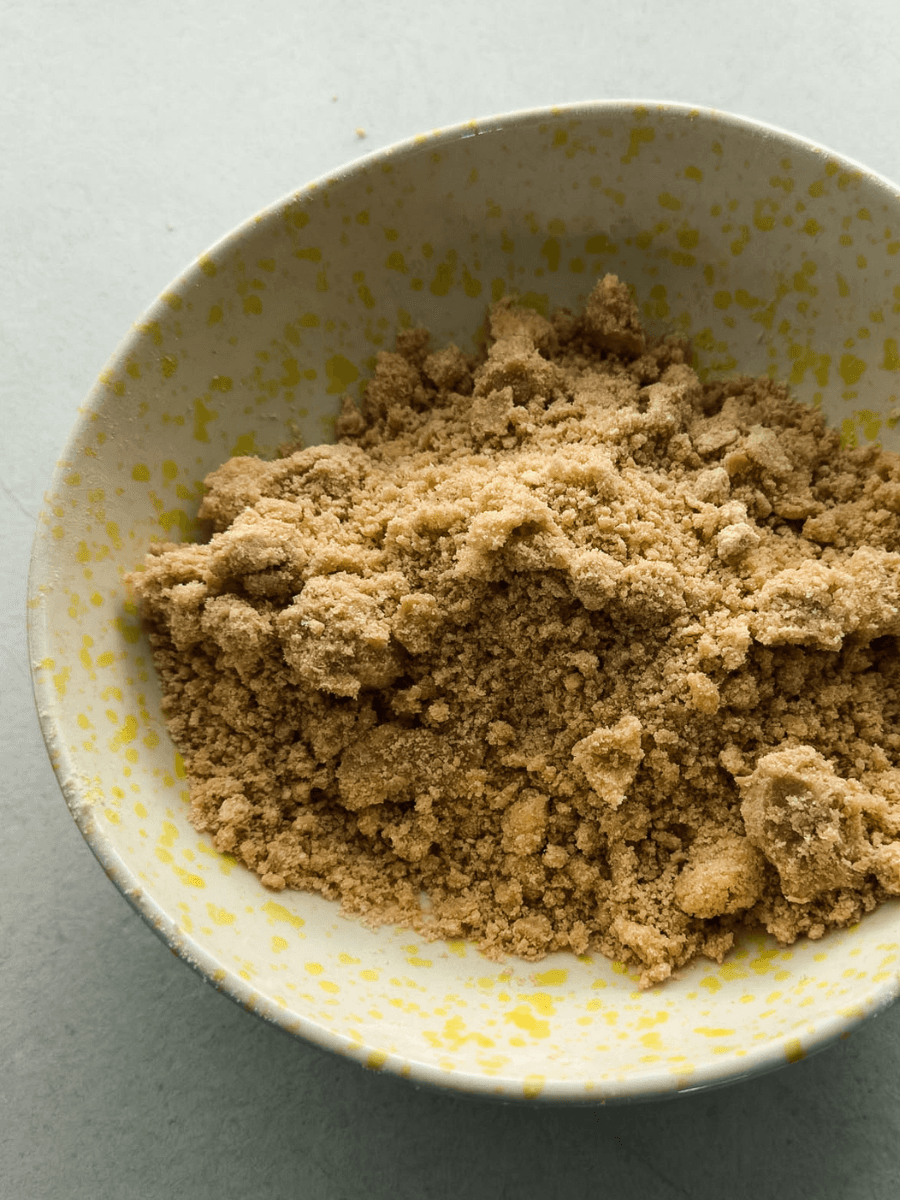 A ceramic bowl filled with a traditional apple crumble topping mixture, showing a sandy, golden-brown texture with both small and large, clumpy butter and flour fragments.