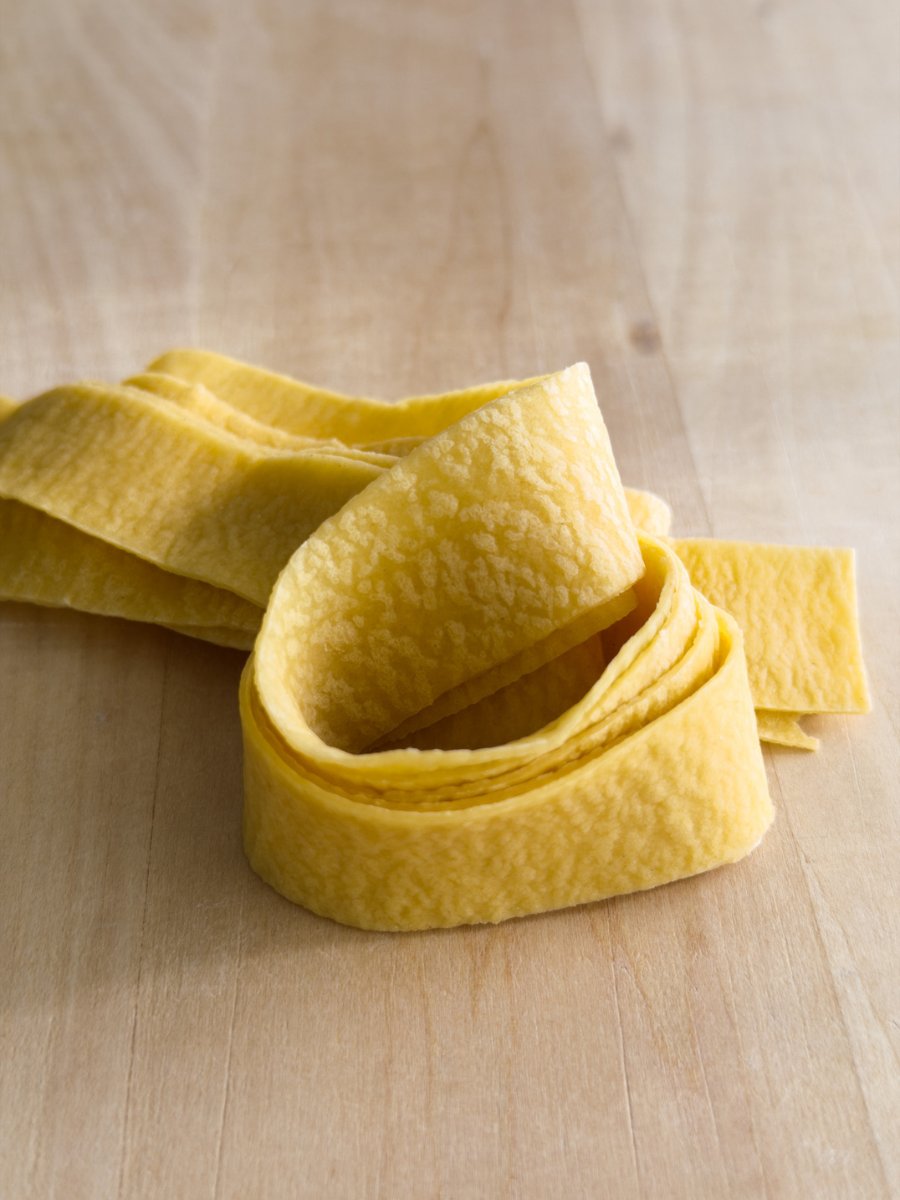 A nest of fresh pappardelle pasta with a rough surface texture sits on a wooden pasta board.