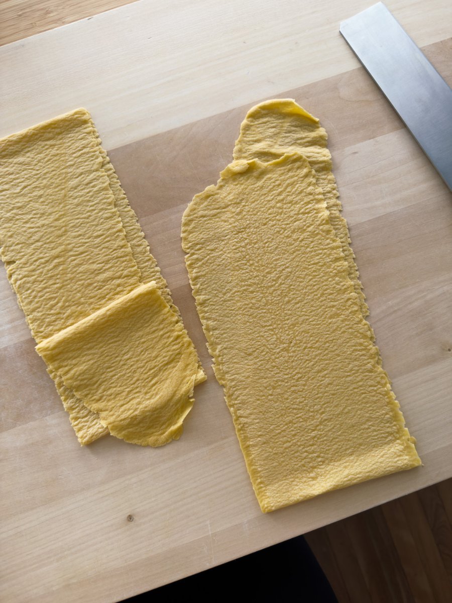 A top down shot of sheets of heavily textured fresh pasta ruvida on a wooden pasta board with a large pasta cutting knife placed nearby.