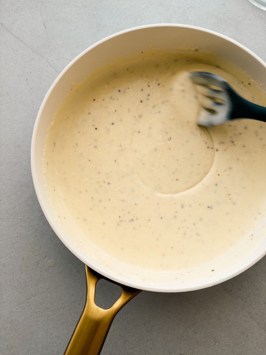A pan of béchamel sauce is stirred by a large spoon, shown from above. 