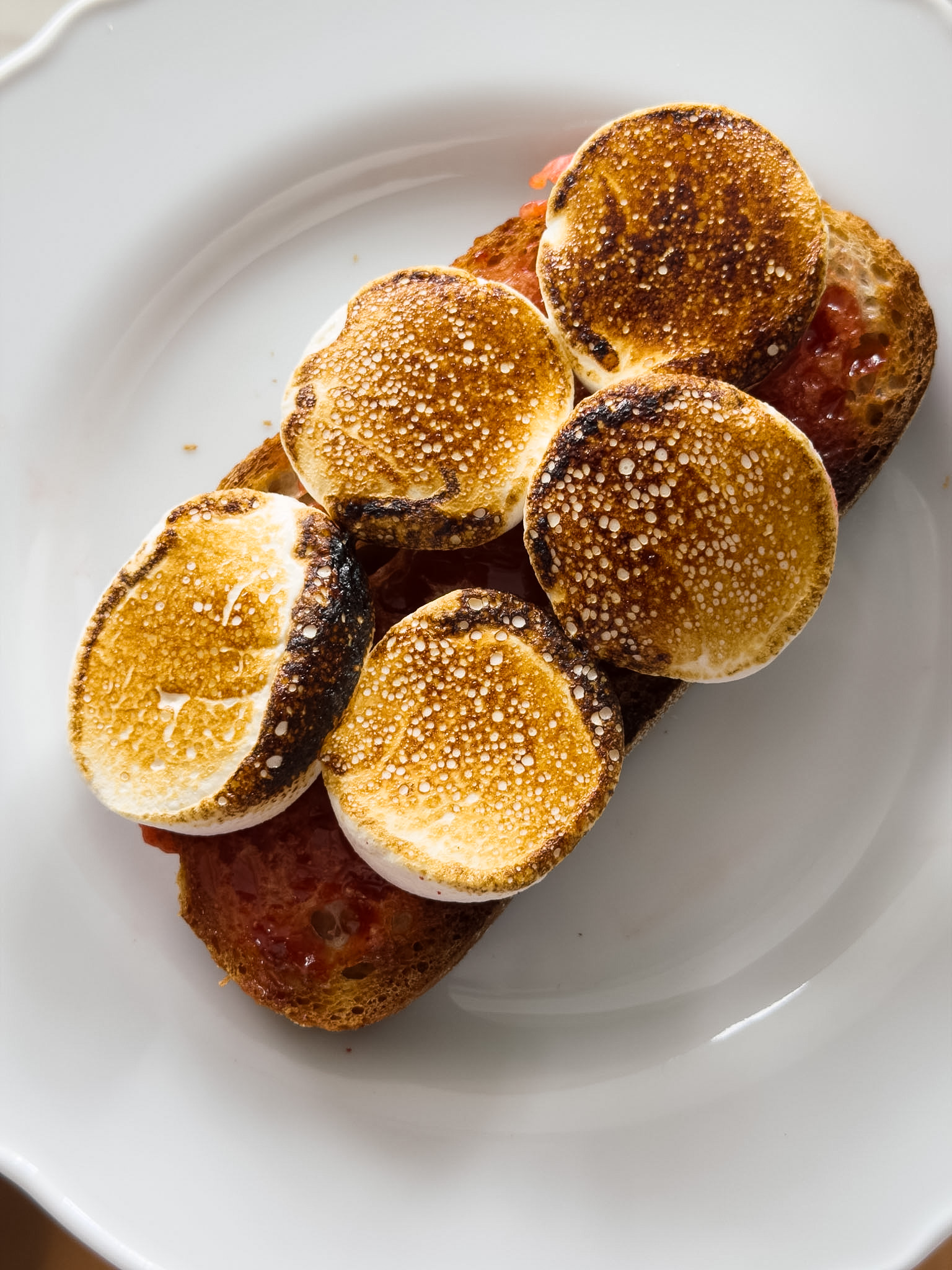 A close up shot of a slice of toast topped with pink strawberry jam and thick slices of marshmallow which have been toasted to perfection.