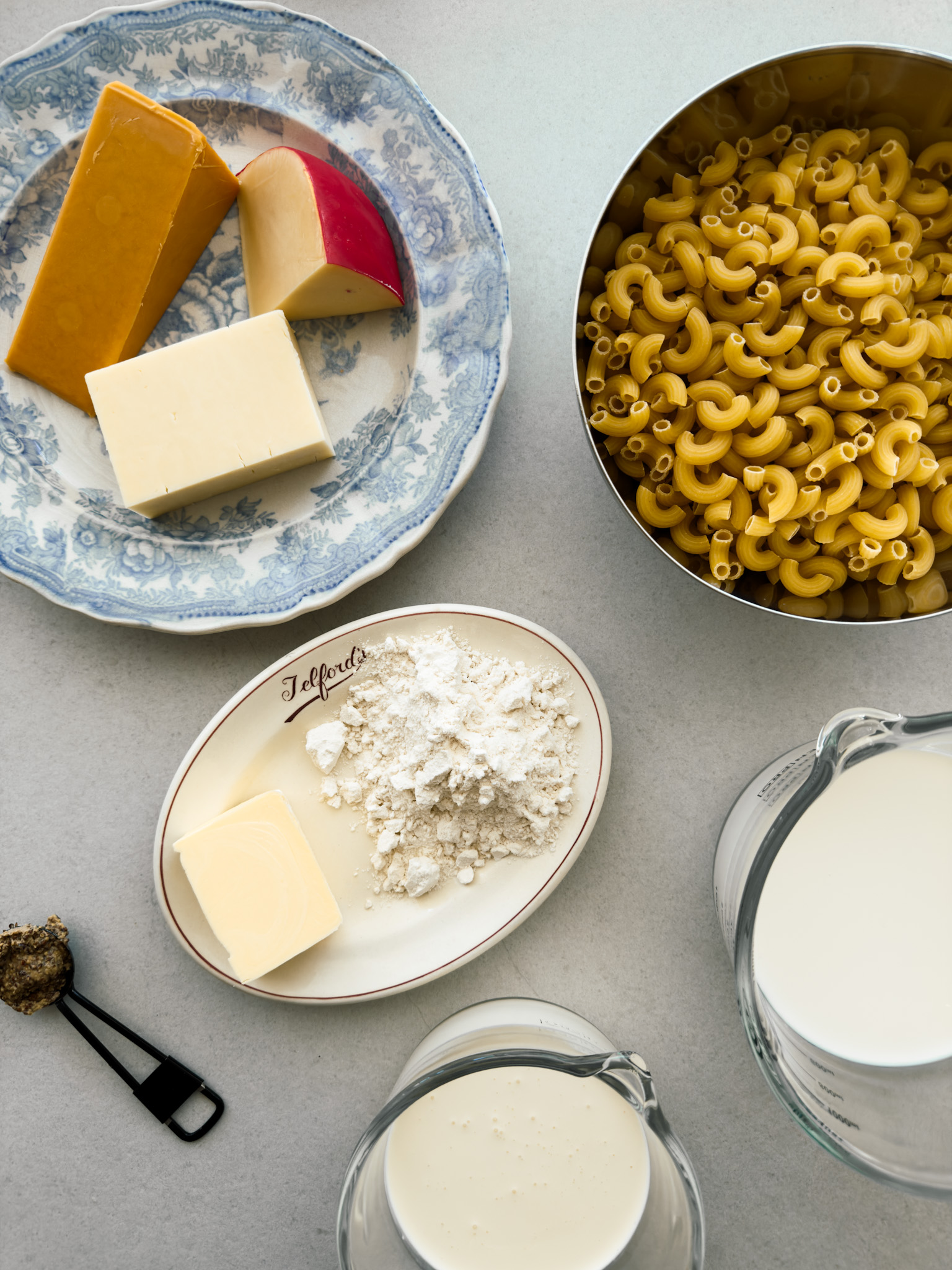 A flat lay of the ingredients needed for the perfect mac and cheese - cheddar, Red Leicester and edam cheese, elbow macaroni, milk, cream, butter, flour and wholegrain mustard. 