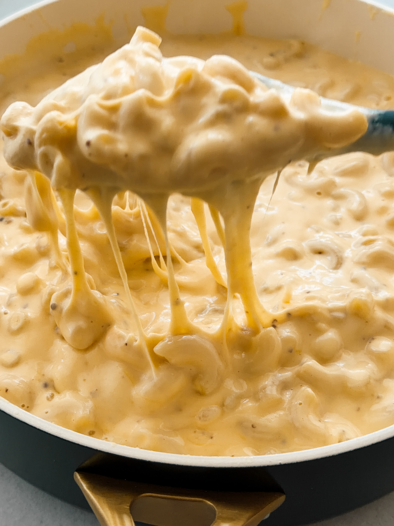 A spoon lifts out some macaroni cheese from a large pan. The sauce is extremely creamy and cheesy, with cheese pulls. 