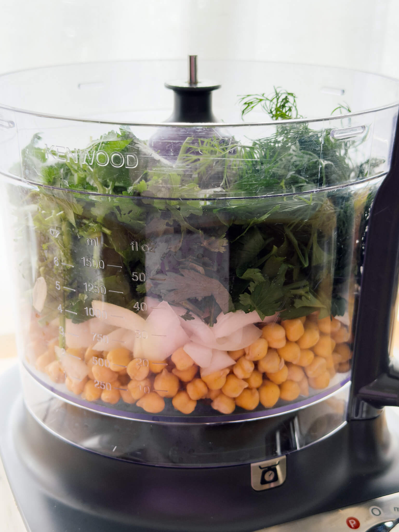 Falafel ingredients shown in a food processor. Layers of soaked chickpeas, onion, spices and green soft herbs. 