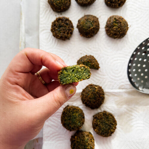 A tray lined with paper towel and freshly fried falafel are shown from above. A hand holds a cut falafel above it, exposing the herby green centre.