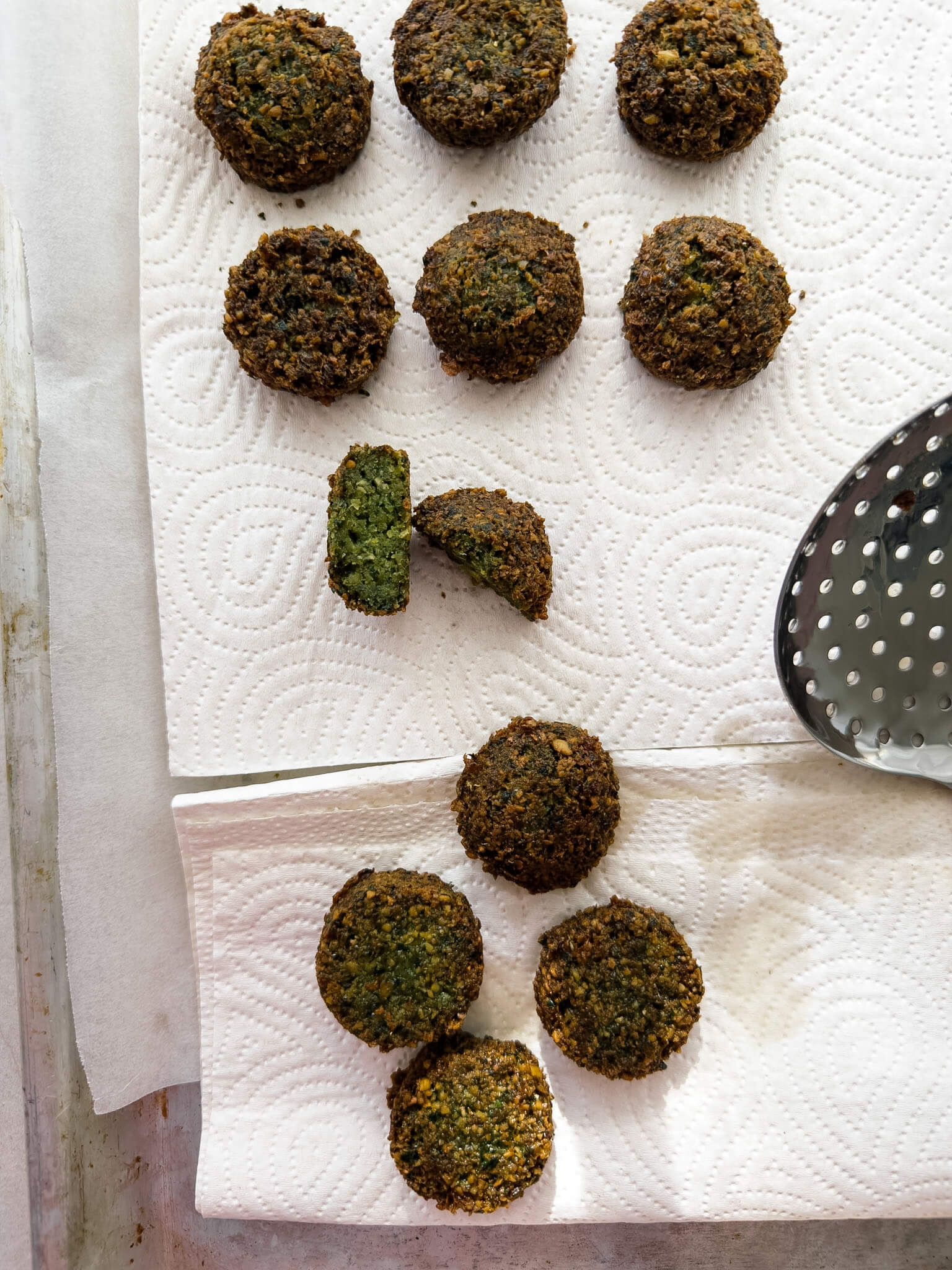 Fried falafel are shown top down on a piece of paper towel. One falafel has been cut in the middle and the mid-section is shown, revealing a crisp exterior and fluffy, vibrant green centre. 